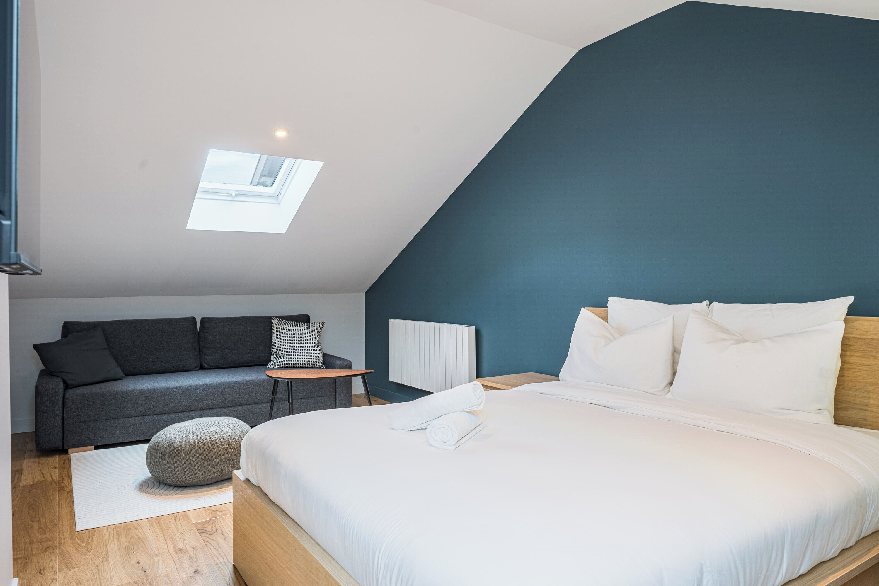 Ein modernes Schlafzimmer mit Schräge, einem Oberlicht, einem Bett mit weißen Leinentüchern, einem grauen Sofa, einem kleinen runden Hocker und einem Holzboden.