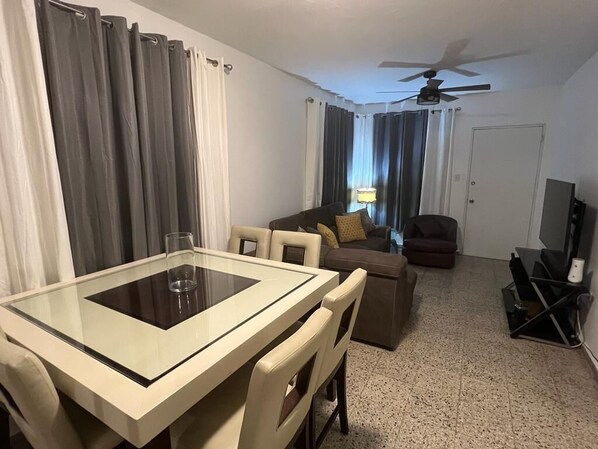 Una habitación de hotel con una mesa de comedor, un sofá, un televisor y un ventilador de techo.