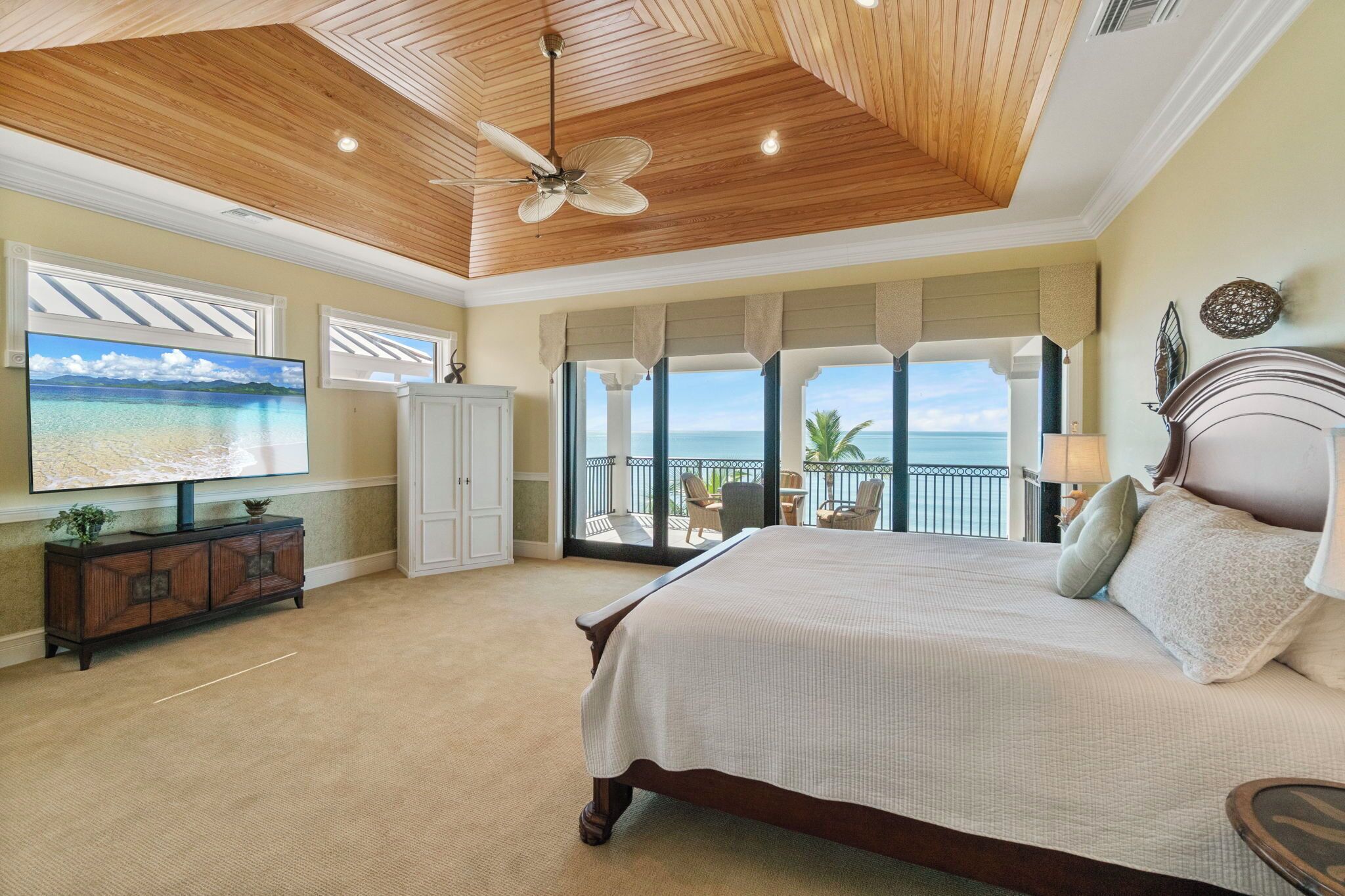 A bedroom with a large bed, a ceiling fan, a TV, and a view of the ocean.