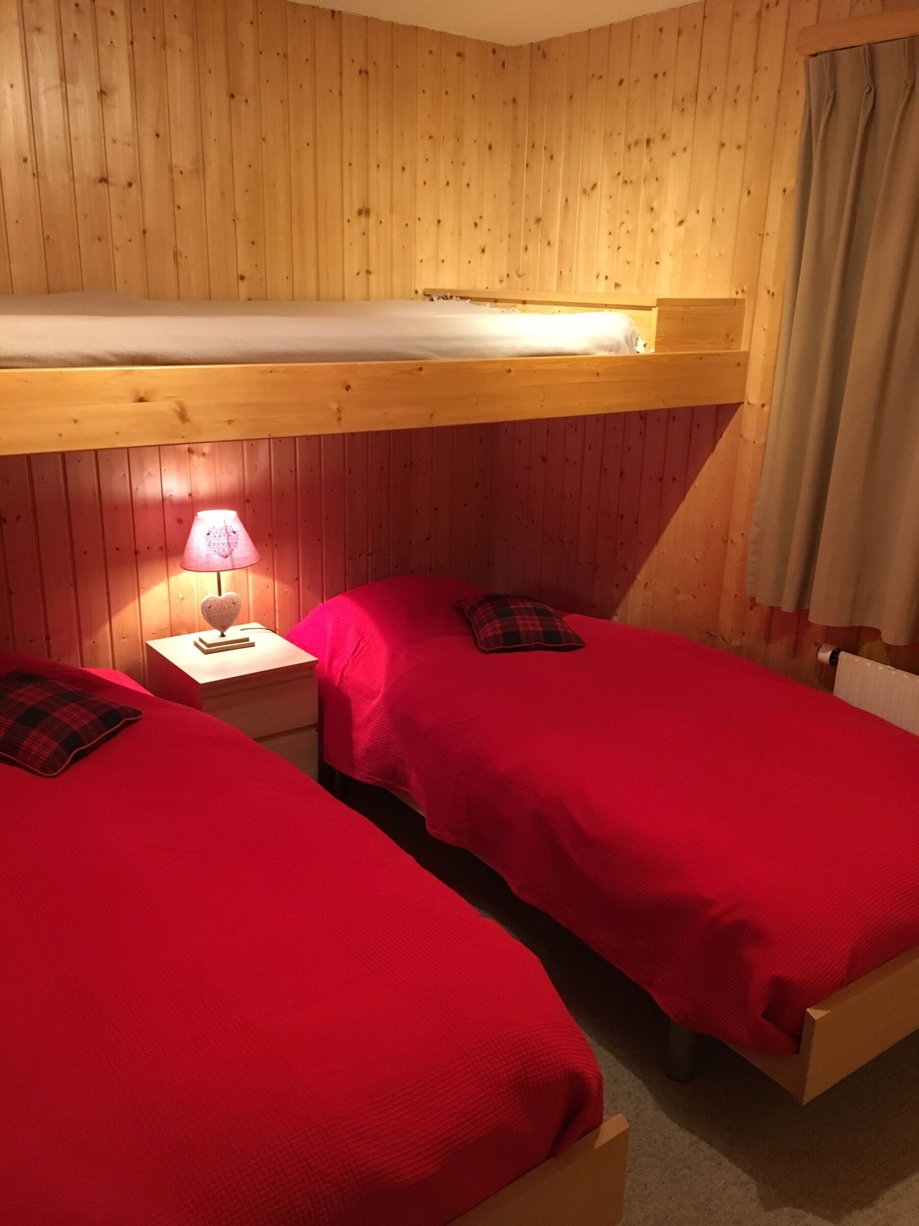 Una stanza con due letti a castello, ognuno con copriletto rosso e un cuscino. Tra i letti, un comodino con una lampada. La stanza presenta pareti in legno e un soffitto spiovente.