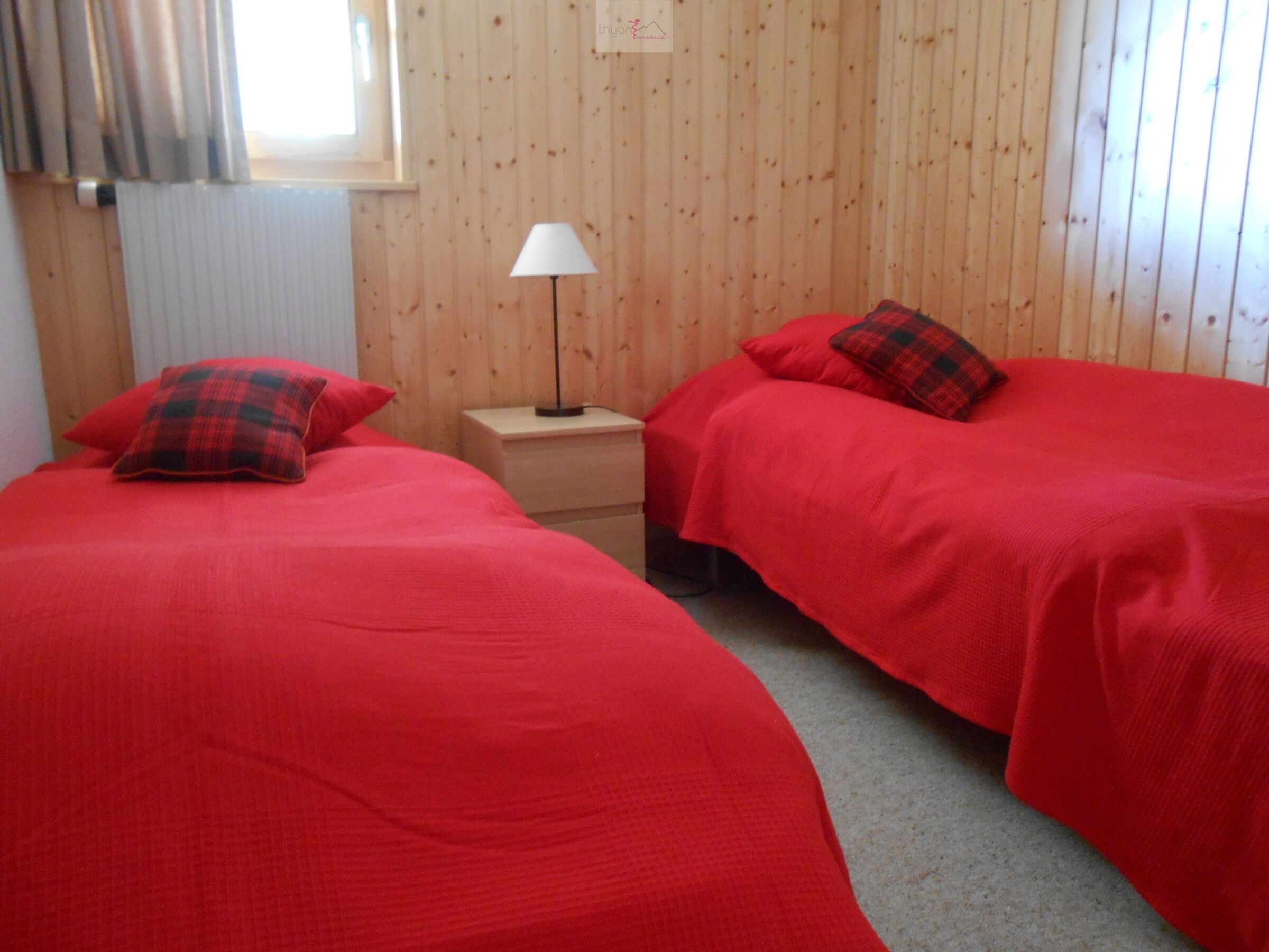A room with two beds, each with red bedding and plaid pillows. There is a bedside table with a lamp between the beds. The walls are wooden paneling.