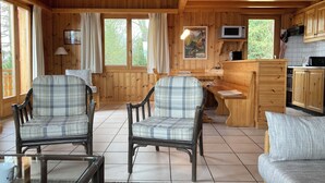 A cozy cabin interior with wooden walls, two plaid chairs, a sofa, a dining table, and a kitchen area with a microwave and cabinets. - Nice new chalet for rent in Les Collons (Vex)