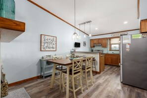 A modern kitchen with wooden cabinets, a stainless steel refrigerator, and a dining area with a wooden table and chairs. - Sea Glass Shack - Condo with Hot Tub, Ocean Views and Great Community Amenities! (Kitty Hawk)