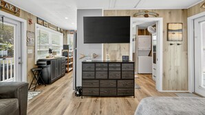 Una sala de estar moderna con un televisor de pantalla plana grande, un mueble de madera oscura y un área de cocina visible a través de una puerta abierta.