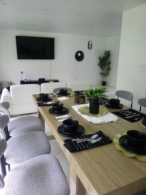 Un coin repas avec une longue table dressée pour le repas, des chaises, un téléviseur et une plante en pot.