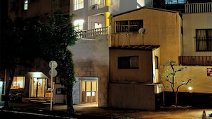 A multi-story building with illuminated windows and balconies, a visible entrance, and a small tree in front. - HOTEL URO - hostel (Nagasaki)