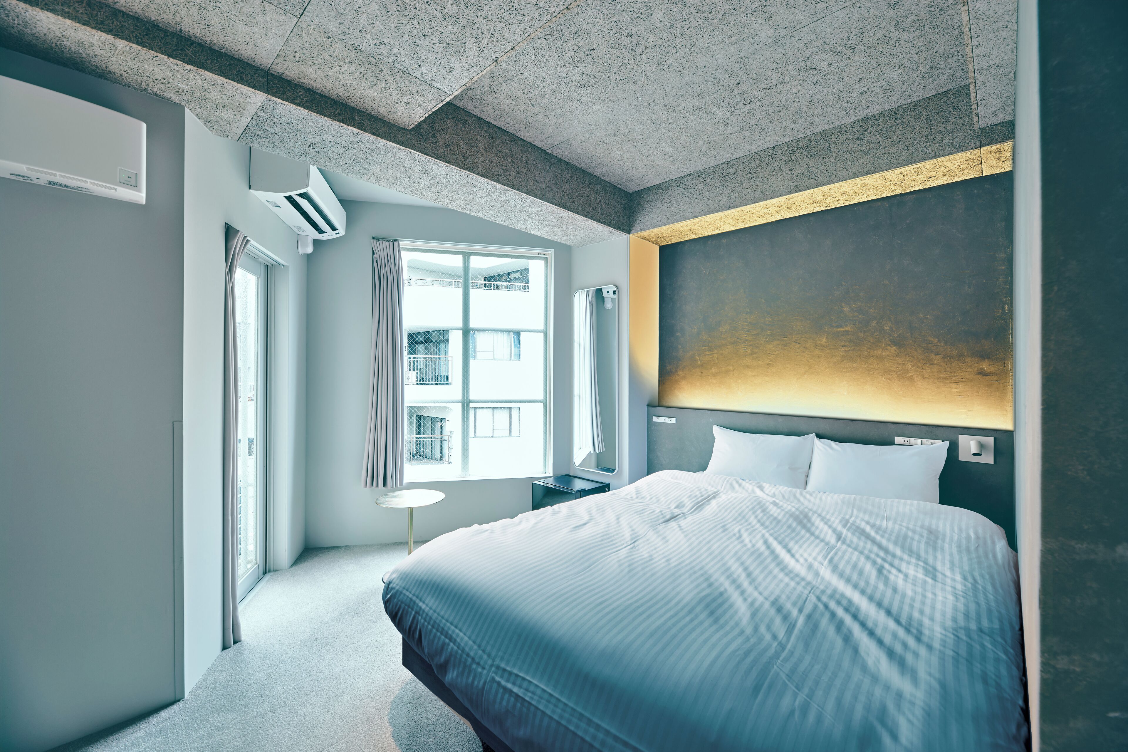 A modern hotel room with a large bed, a small bedside table, a window with curtains, and an air conditioning unit.
