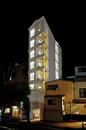 A multi-story building with illuminated windows and balconies, a visible entrance, and a small tree in front. - HOTEL URO - hostel (Nagasaki)
