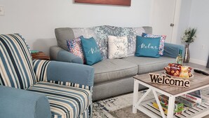 Un salon avec un canapé à rayures, un divan gris et une table basse sur laquelle est posée une pancarte indiquant « Bienvenue ».