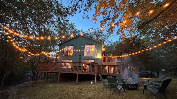 Une maison à deux étages avec une terrasse, entourée d’arbres et illuminée de guirlandes lumineuses.