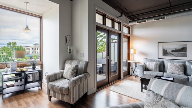 Un salon moderne avec un canapé, un fauteuil et une table basse. Une grande fenêtre offre une vue sur les immeubles et les arbres.