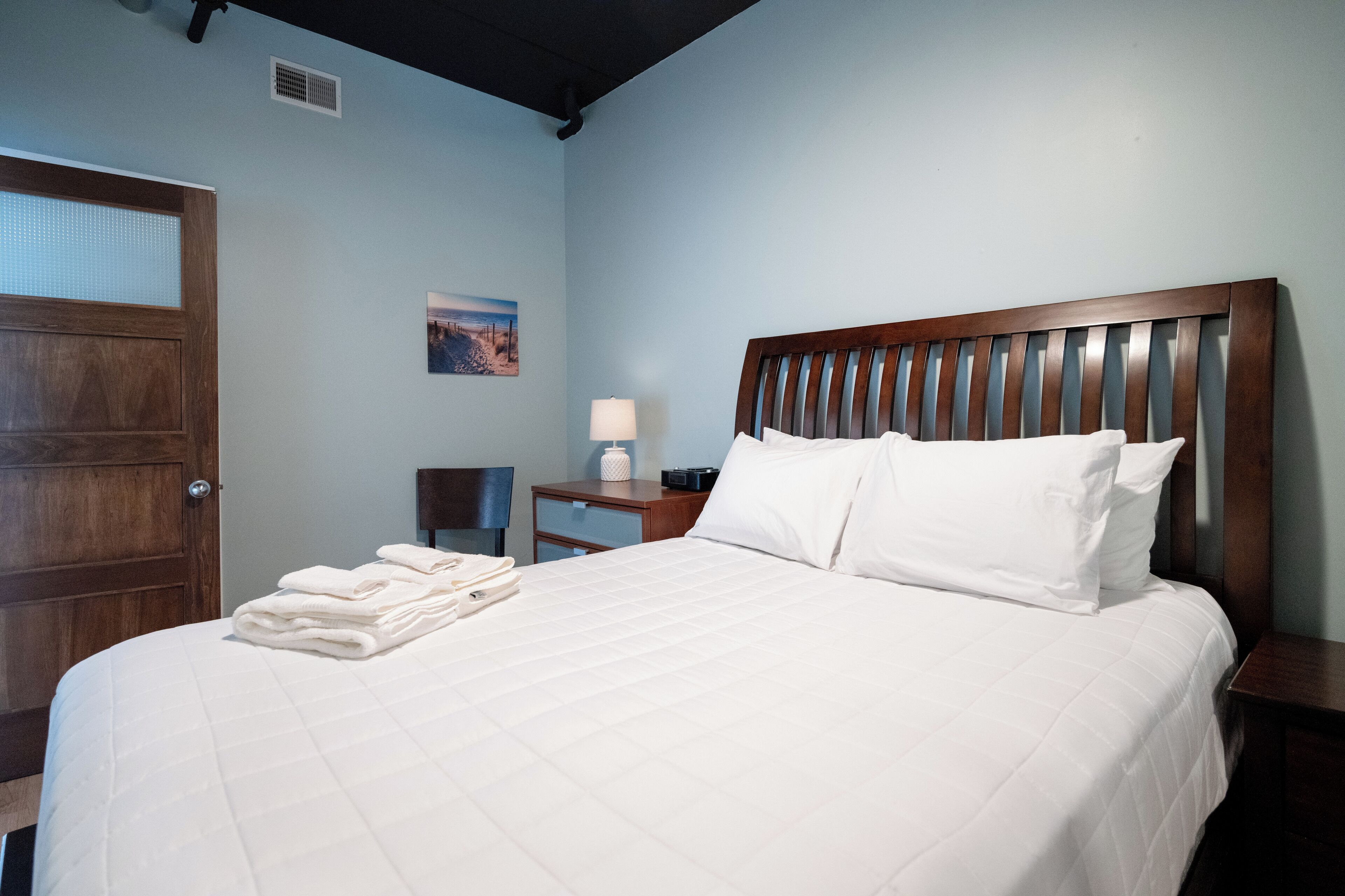 A neatly made bed with white linens, a wooden headboard, and a nightstand with a lamp and a small table.