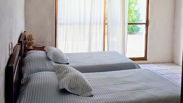 A bedroom with two beds, a wooden headboard, a window with curtains, and a view of greenery outside.
