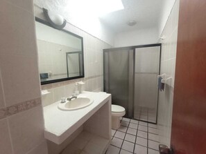 Une salle de bain avec un lavabo blanc, un miroir et des toilettes. On y trouve une douche en verre et une porte sur la droite.