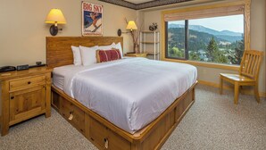 Habitació d'hotel amb un llit gran, un capçal de llit de fusta, dues làmpades de nit, una taula de nit, una cadira, una finestra amb vistes a les muntanyes i un cartell a la paret.