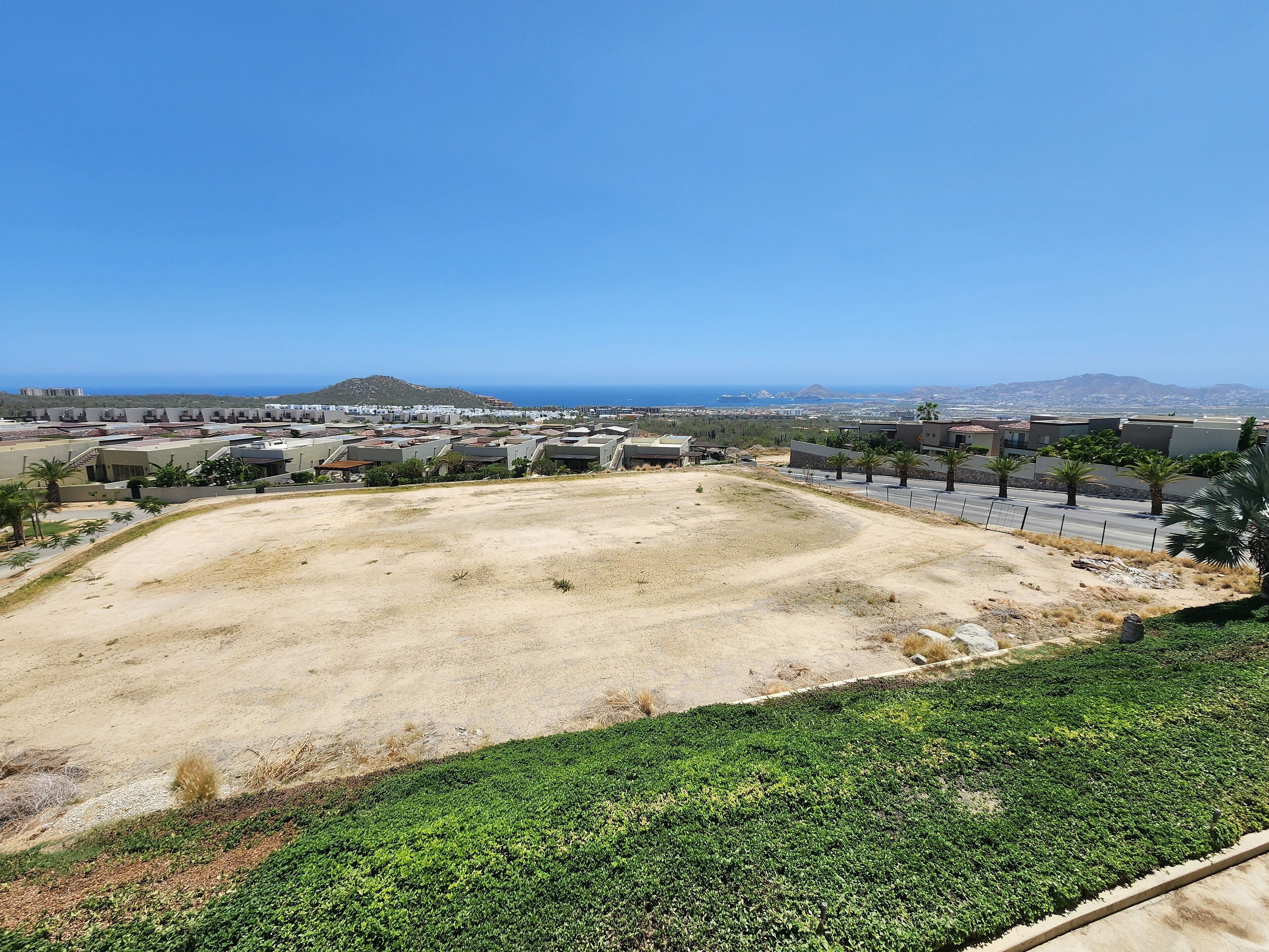 A residential area with a large open space, a road with palm trees, and buildings in the background.