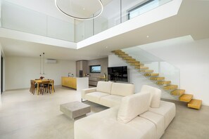 A modern interior with a high ceiling, a staircase with glass railings, a dining area with a table and chairs, and a living room with a sofa and a flat-screen TV.