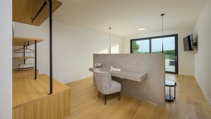 A modern, minimalist room with a wooden floor, a grey upholstered chair, a built-in desk with a book and a container, and a large window offering a view of greenery.
