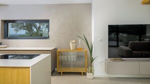 A modern living room with a large window, a glass cabinet with wine glasses, a potted plant, and a flat-screen TV.