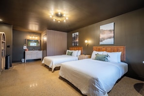 Quarto de hotel com duas camas, uma penteadeira com espelho e uma janela grande com vista para árvores.