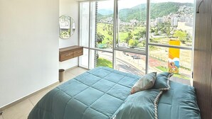 A bedroom with a large bed, a wooden headboard, a window with a view of a city and mountains, and a small bedside table. - 3 bedroom family apartment in Acapulco, a few steps from the beach. (Acapulco de Juárez)