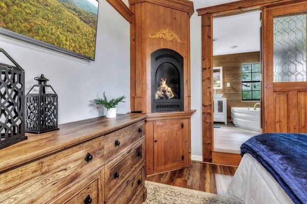 A wooden dresser with drawers, a fireplace, a bed with a blue blanket, and a bathroom with a bathtub and a window.