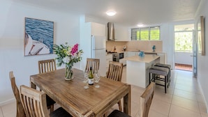 A dining area with a wooden table and chairs, a vase with flowers, and a kitchen in the background.