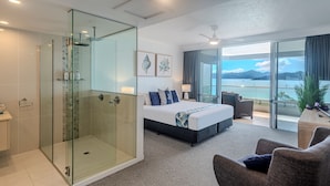 A modern hotel room with a glass-enclosed shower, a large bed with a view of the sea, and a balcony with a view of the mountains.