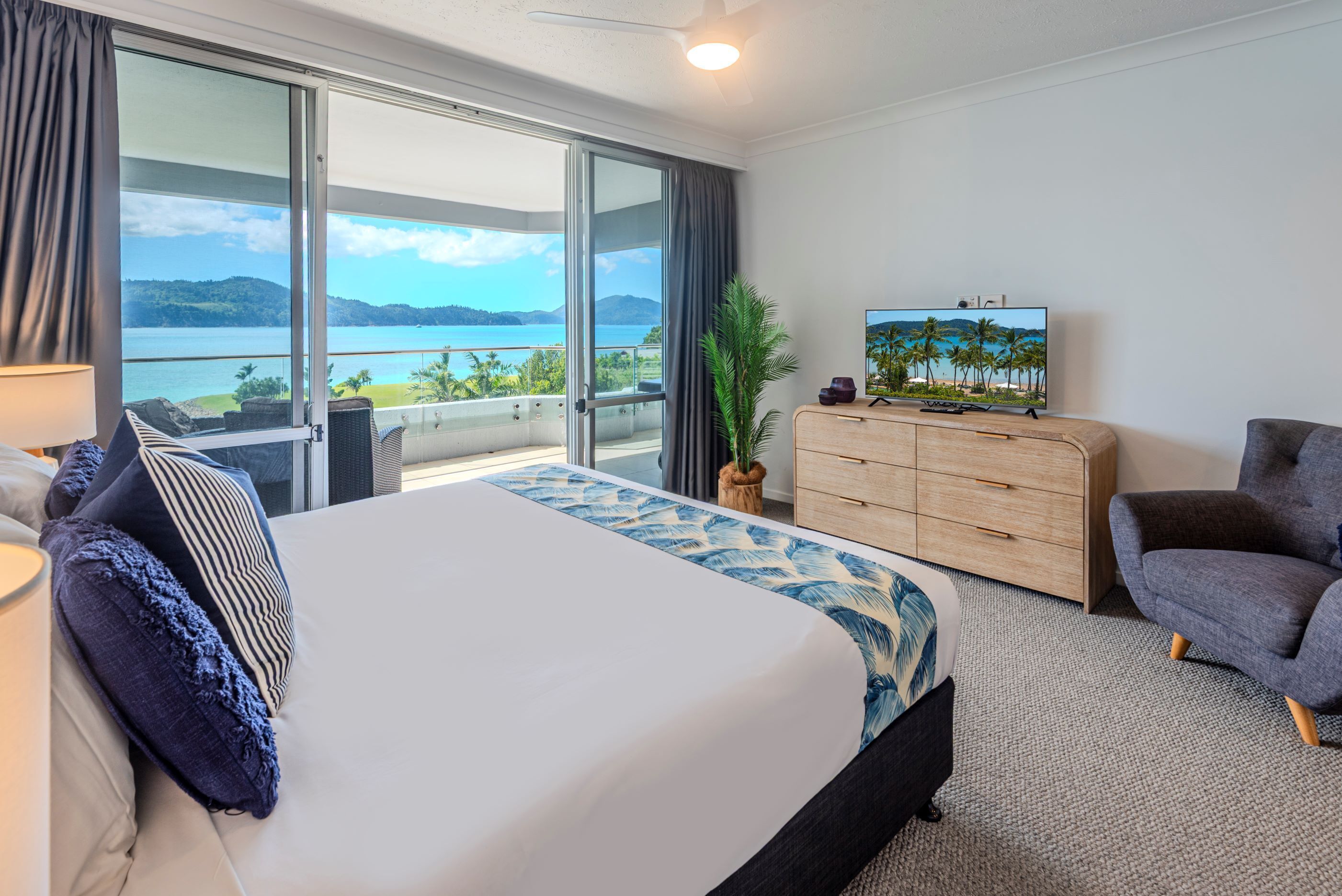 A hotel room with a large bed, a TV, a wooden dresser, a grey armchair, and a view of the sea.
