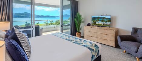 A hotel room with a large bed, a TV, a wooden dresser, a grey armchair, and a view of the sea.
