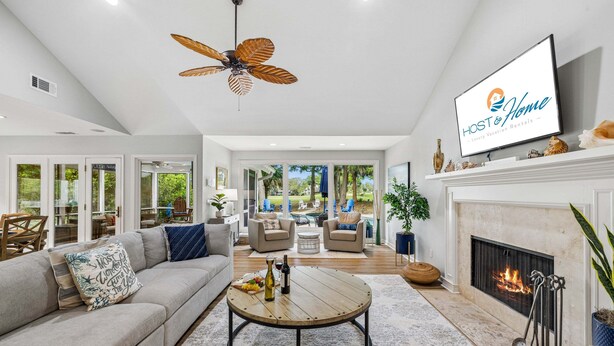 A spacious living room with a fireplace, a ceiling fan, and a television displaying 'HOST & Home'. - 22 Heath Drive @ Palmetto Dunes | Private Pool | Golf Course View (Hilton Head Island)