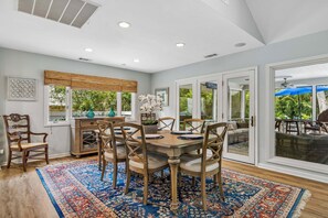 A dining area with a wooden table and chairs, a patterned rug, and a view of the outdoors through large windows.