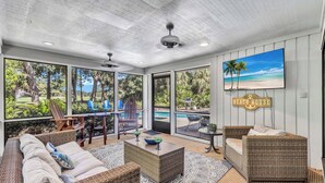 A spacious room with a large window overlooking a pool and lush greenery. There is a wicker sofa, a small table with a bowl and glasses, and a sign reading "BEACH HOUSE". - 22 Heath Drive @ Palmetto Dunes | Private Pool | Golf Course View (Hilton Head Island)