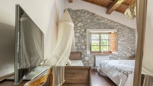 A bedroom with a stone wall, a bed with a canopy, a dresser, a mirror, and a window with shutters. - Llanes House with Garden - La Casa del Güelu (Hontoria)