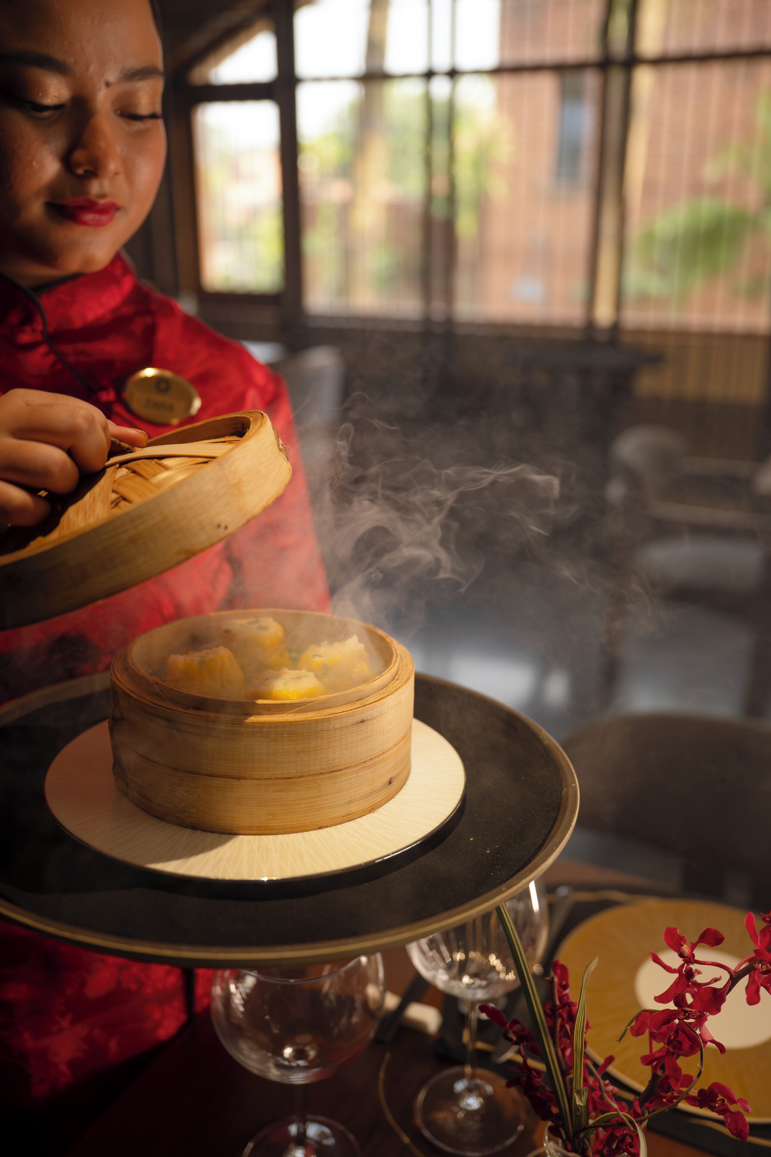 A person in a red outfit serving food from a bamboo steamer, with steam rising, on a table with a wine glass and a bowl.