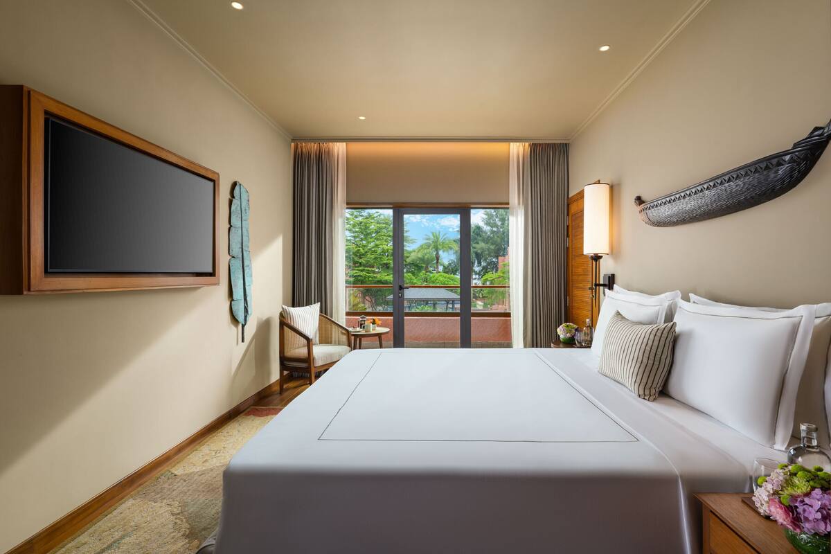 Quarto de hotel com cama grande, uma cadeira, uma mesinha e vista para o verde lá fora.