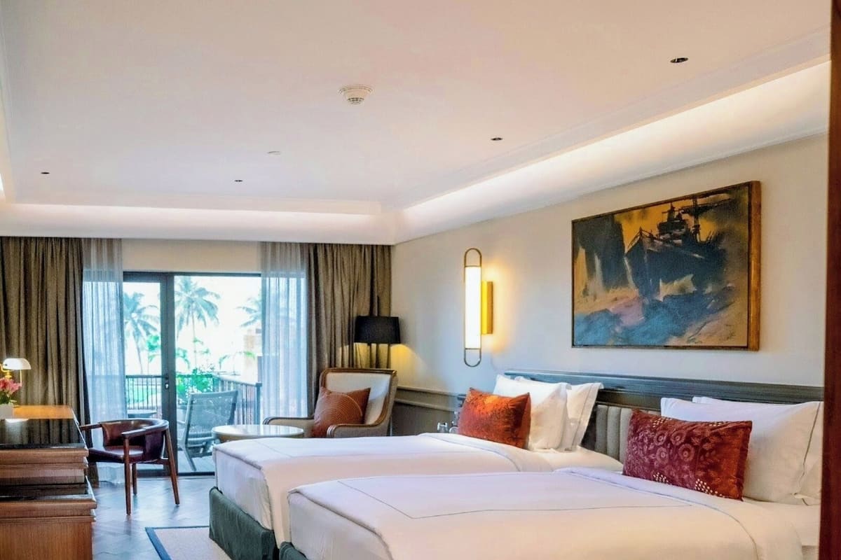 Quarto de hotel com duas camas, uma escrivaninha, uma cadeira, um abajur e um quadro na parede.