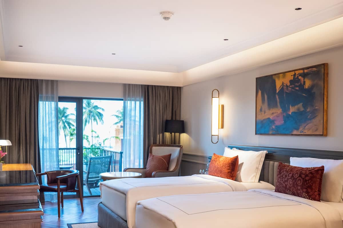 Quarto de hotel com duas camas, uma escrivaninha, uma cadeira e varanda com vista para palmeiras.