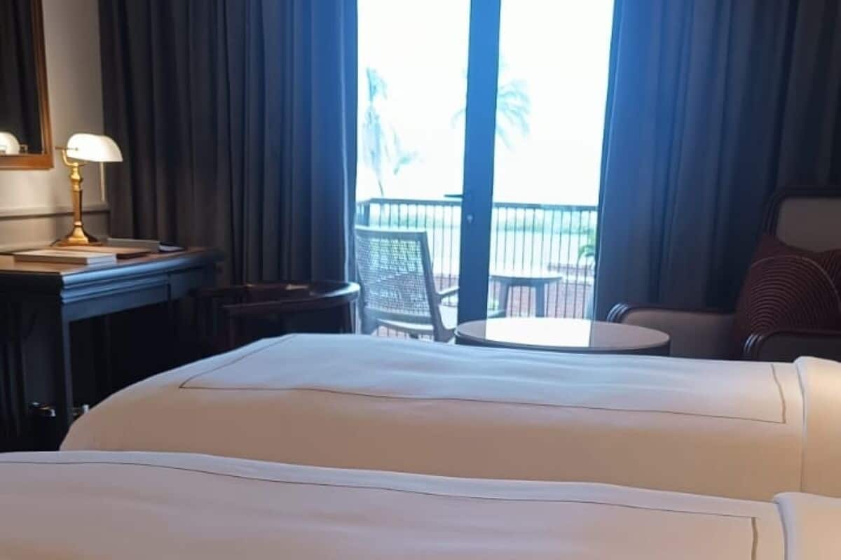 Quarto de hotel com uma cama grande, uma escrivaninha com luminária, uma cadeira e vista para o exterior através de uma janela.