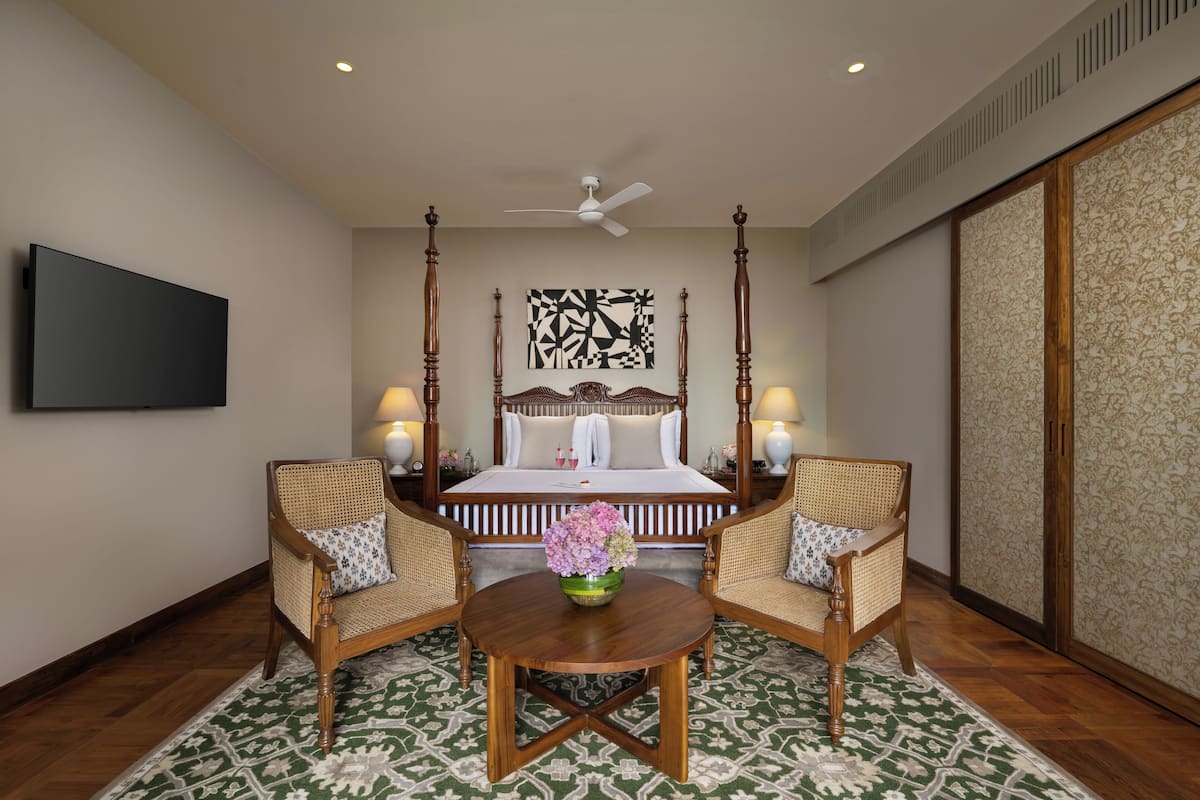 Quarto com cama, duas cadeiras, mesa, ventilador de teto e TV na parede.