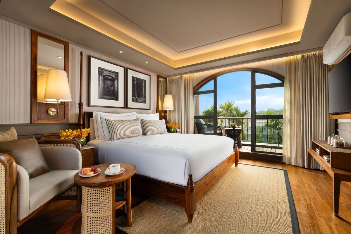 Quarto de hotel com uma cama grande, um sofá, uma mesa pequena com uma chávena e um prato, e uma varanda com vista.