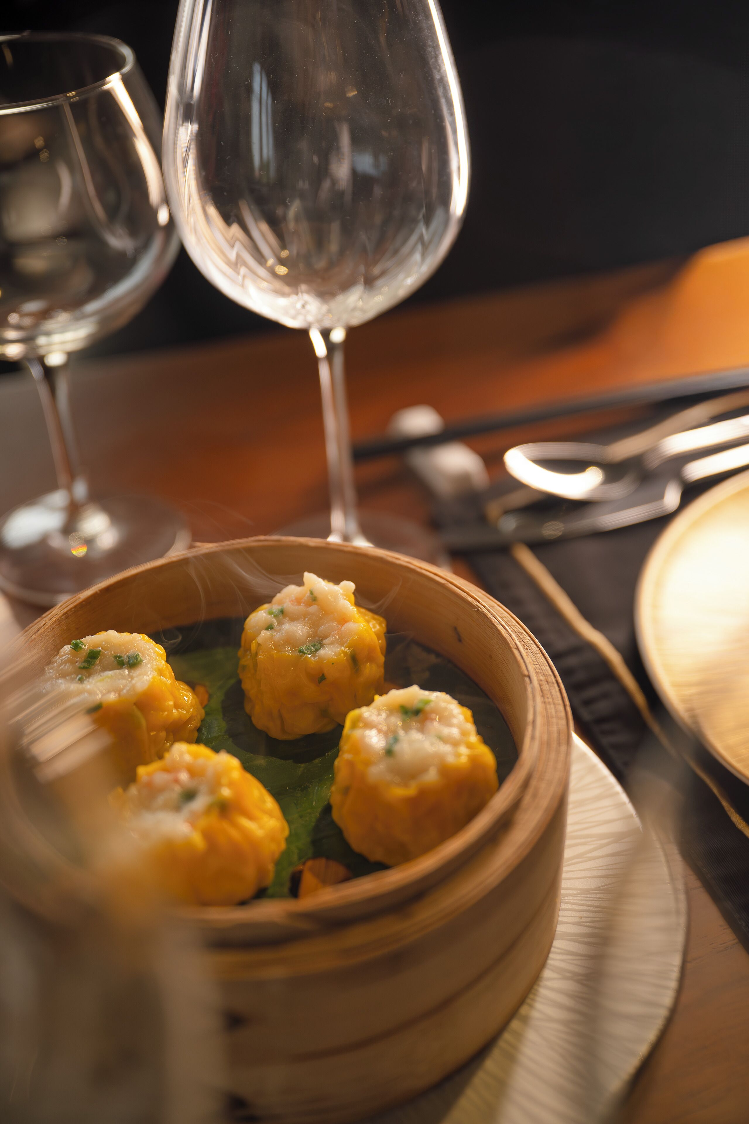 Un tazón de madera con dumplings en un plato, acompañado por una copa de vino y cubiertos sobre una mesa.