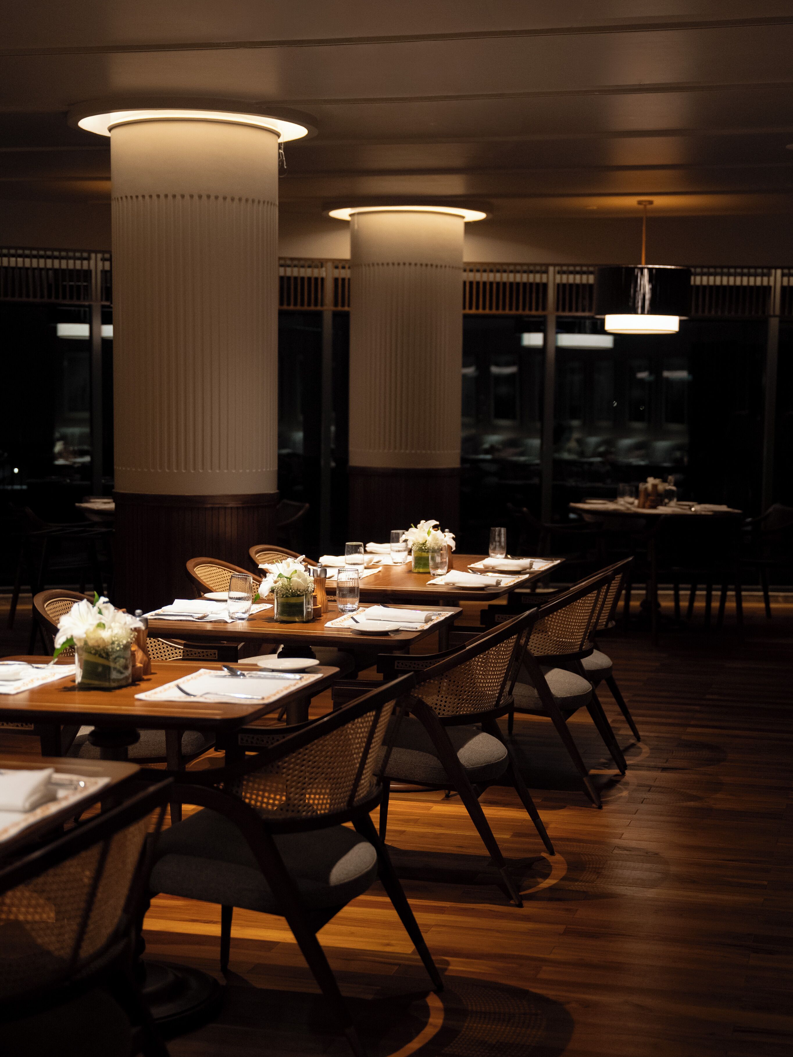 Un ristorante scarsamente illuminato con tavoli e sedie in legno, ampi lampadari cilindrici e vista all'esterno tramite ampie finestre.
