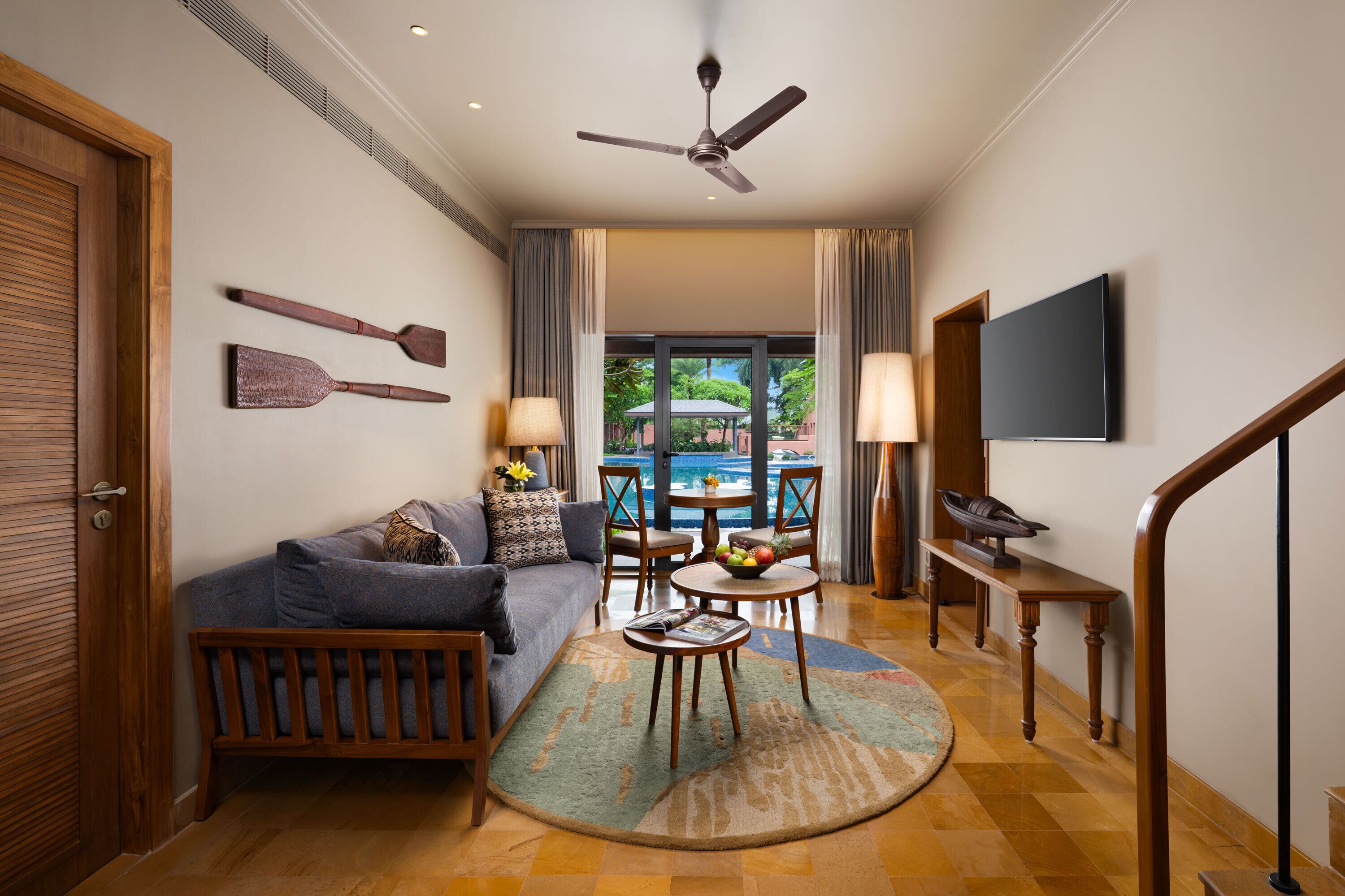 A living room with a sofa, coffee table, and a TV. There is a ceiling fan, a window with curtains, and a view of a pool outside.
