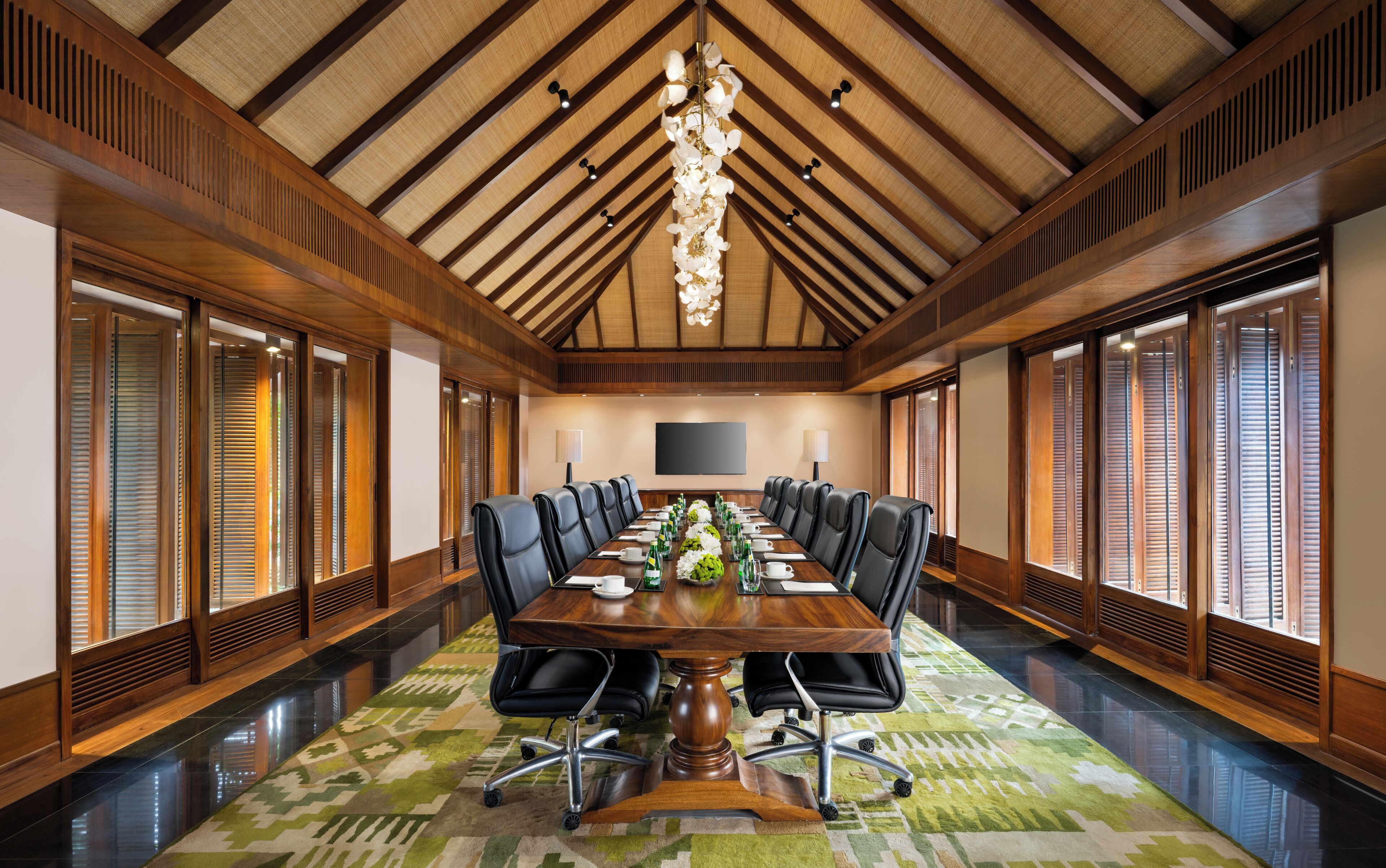 A conference room with a long table, black chairs, a chandelier, and large windows with blinds.