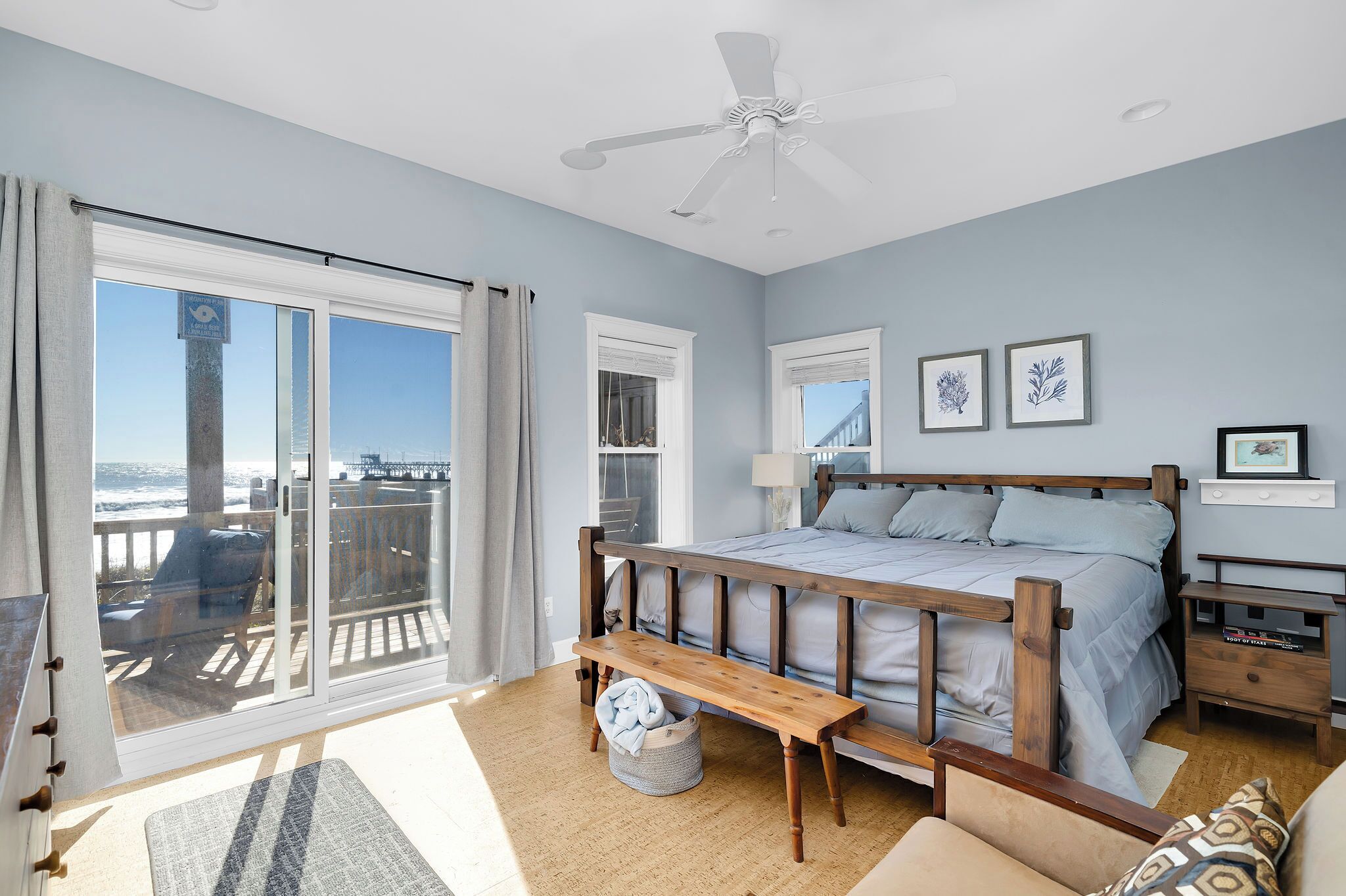 A bedroom with a large bed, a ceiling fan, a view of the ocean through a sliding door, and a small bedside table.