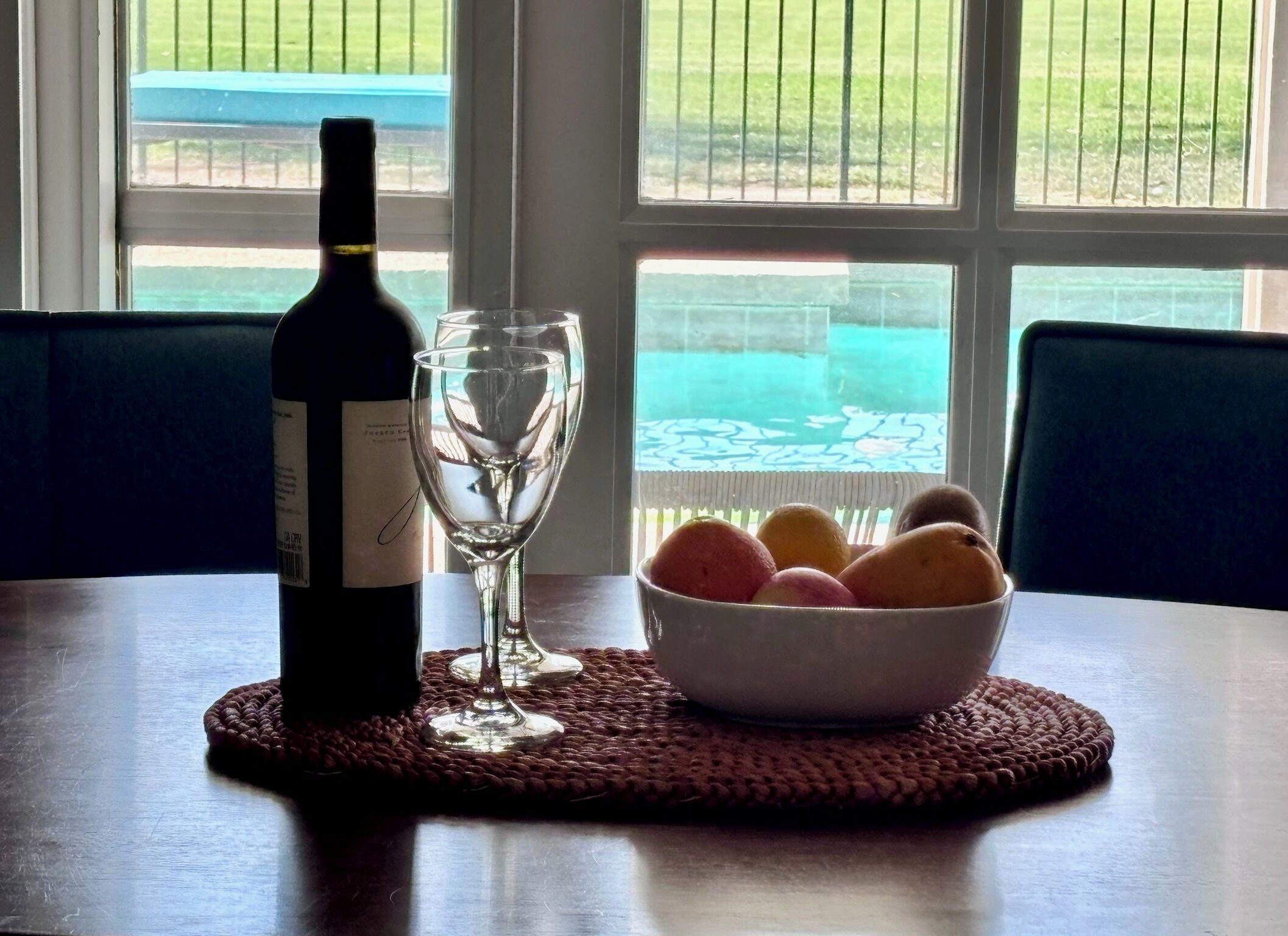 Una botella de vino, una copa vacía y un cuenco de fruta sobre una mesa junto a una ventana con vistas a una piscina y un jardín.