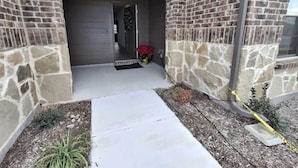 A building entrance with a stone facade, a concrete pathway, and a small garden area with plants and a potted plant.