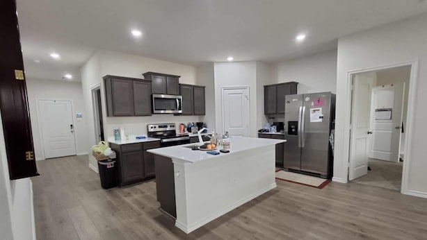 A modern kitchen with dark cabinets, a white island, stainless steel appliances, and a wooden floor.