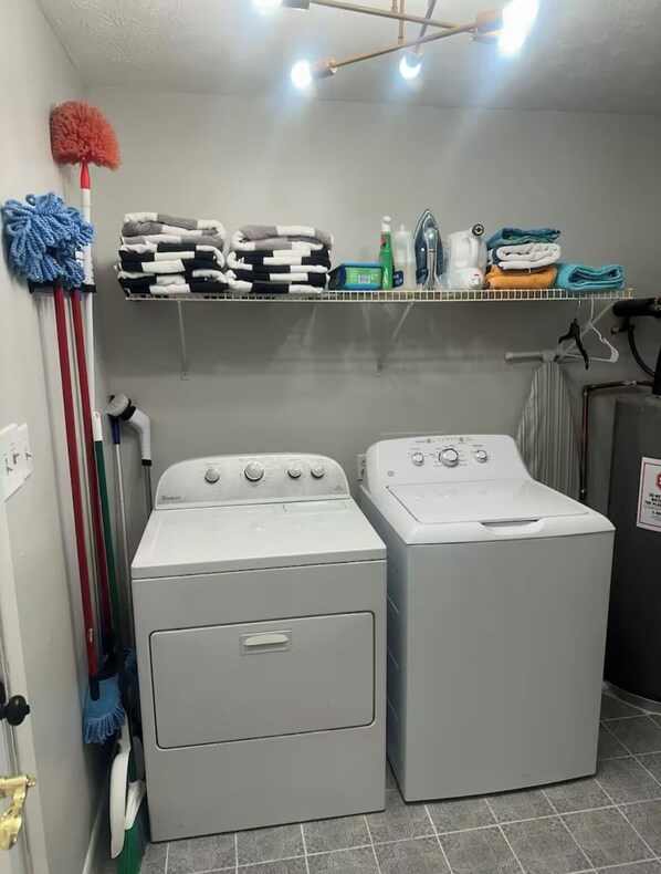 A laundry room with two front-loading washing machines, a shelf with folded towels and cleaning supplies, and a mop on the wall.